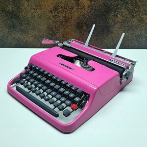 May include: A pink vintage typewriter with a black keyboard and silver accents. The typewriter is on a white surface.