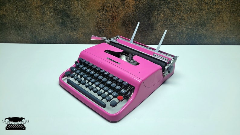 May include: A pink vintage typewriter with a black keyboard and silver accents. The typewriter is on a white surface.