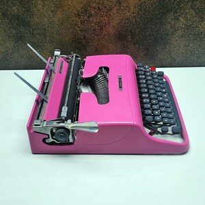 May include: A pink vintage typewriter with a black keyboard and silver keys. The typewriter has a carriage return lever and a paper roller.