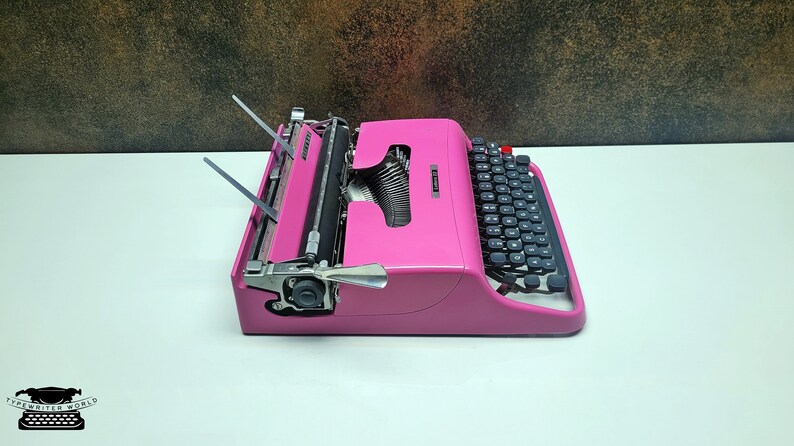 May include: A pink vintage typewriter with a black keyboard and silver keys. The typewriter has a carriage return lever and a paper roller.