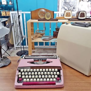 May include: A pink Brother typewriter with a wooden desk in the background. The typewriter has a black carriage return lever and a red tab key. The desk has a variety of vintage items on it, including clocks, a coffee grinder, and a wooden boat.