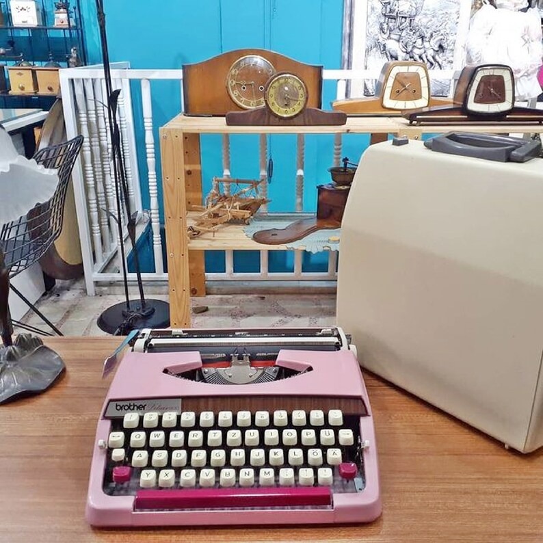 May include: A pink Brother typewriter with a wooden desk in the background. The typewriter has a black carriage return lever and a red tab key. The desk has a variety of vintage items on it, including clocks, a coffee grinder, and a wooden boat.