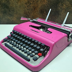 May include: A pink vintage typewriter with black keys and a silver carriage return lever. The typewriter has a white paper holder and a black lever on the right side.