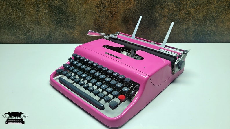 May include: A pink vintage typewriter with black keys and a silver carriage return lever. The typewriter has a white paper holder and a black lever on the right side.