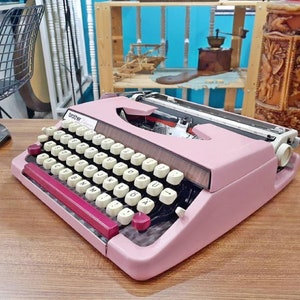 May include: A pink Brother typewriter with a black and brown keyboard and a red return bar. The typewriter has a white label that says "Brother Deluxe".