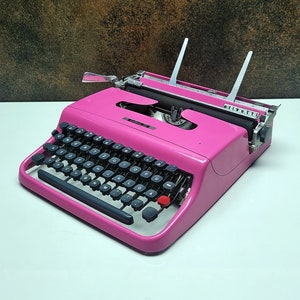 May include: A pink vintage Olivetti Lettera 32 typewriter with a black keyboard and a red return key.