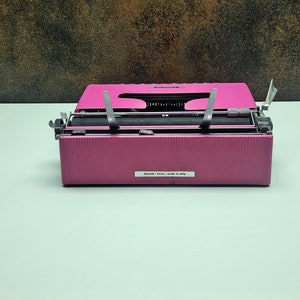 May include: A pink vintage typewriter with a black ribbon and silver keys. The typewriter has a label that reads "Made in Italy".
