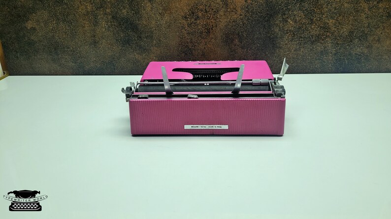 May include: A pink vintage typewriter with a black ribbon and silver keys. The typewriter has a label that reads "Made in Italy".