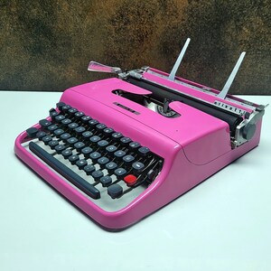 May include: A pink vintage typewriter with a black keyboard and silver accents. The typewriter is on a white surface.