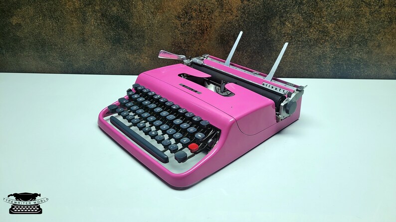 May include: A pink vintage typewriter with a black keyboard and silver accents. The typewriter is on a white surface.