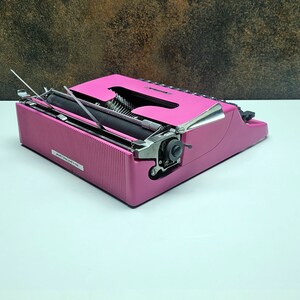 May include: A pink vintage typewriter with a silver carriage return lever and a black ribbon. The typewriter is open and ready to type.