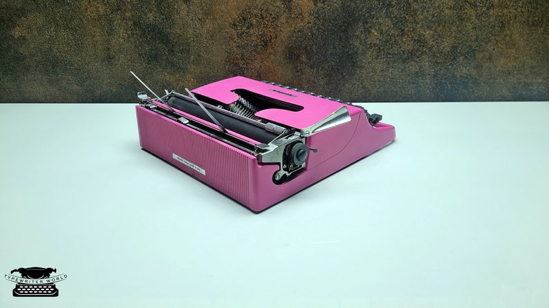 May include: A pink vintage typewriter with a silver carriage return lever and a black ribbon. The typewriter is open and ready to type.