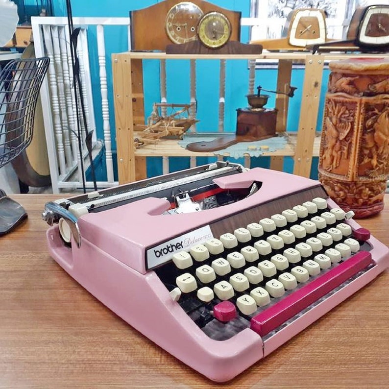 May include: A pink Brother Deluxe typewriter with a black and white keyboard and a burgundy carriage return lever. The typewriter is sitting on a wooden surface.
