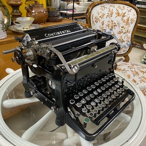 May include: A vintage black typewriter with the word "CONTINENTAL" printed on the front. The typewriter is sitting on a white table with a glass top.
