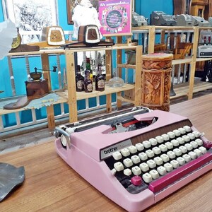 May include: A pink Brother typewriter with a black keyboard and a red carriage return lever. The typewriter is sitting on a wooden table.