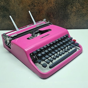 May include: A pink vintage typewriter with black keys and a silver carriage return lever. The typewriter has a white paper holder and a red button on the side.