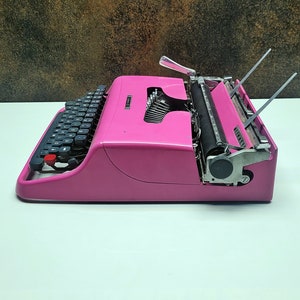 May include: A pink vintage typewriter with a black keyboard and a silver carriage return lever. The typewriter is on a white surface.
