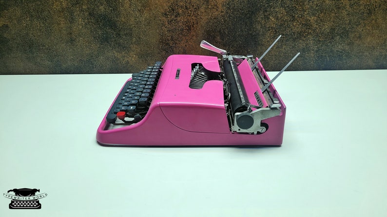 May include: A pink vintage typewriter with a black keyboard and a silver carriage return lever. The typewriter is on a white surface.