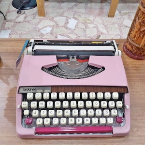 May include: A pink Brother Deluxe typewriter with a white and black keyboard. The keys have letters in both English and German.