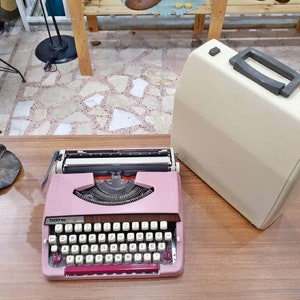 May include: A pink Brother typewriter with a white case. The typewriter has a standard keyboard layout with keys for letters, numbers, and punctuation. The case has a handle on the top.