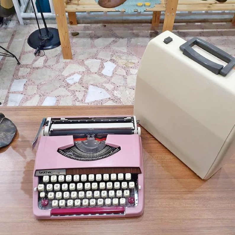 May include: A pink Brother typewriter with a white case. The typewriter has a standard keyboard layout with keys for letters, numbers, and punctuation. The case has a handle on the top.