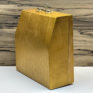 May include: A golden-toned wooden box with a hinged lid and metal clasp. The box has a rectangular shape with angled sides, and a light-colored wood grain is visible. The box is sitting on a white surface.