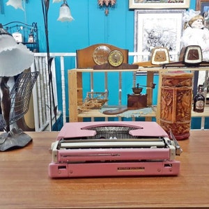 May include: A pink vintage typewriter with a white keyboard and the text "Brother  Made in Japan" on the bottom. The typewriter is sitting on a wooden table.