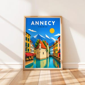 Puede incluir: Un póster de viaje enmarcado con una ilustración colorida de Annecy, Francia. La imagen muestra un canal entre edificios de colores, una estructura histórica central con techo rojo y montañas nevadas bajo un sol amarillo brillante.