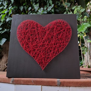 May include: A red string art heart on a dark gray wooden background.