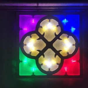 May include: A decorative wall art piece with a four-leaf clover design. The clover is made of white wood and has a yellow light inside. The background is made of four squares of colored glass: purple, blue, green, and red.