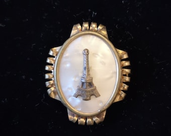 May include: A gold-tone brooch with a white cameo featuring a detailed depiction of the Eiffel Tower.