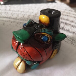 May include: A ceramic pipe shaped like a stylised animal head, featuring a glossy finish in vibrant colours including red, green, yellow, and blue. The design includes a prominent snout, textured details, and a cylindrical bowl extending from the top.