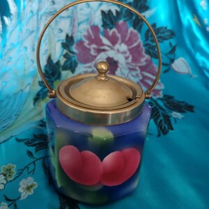 May include: A blue and white ceramic jar with a brass lid and handle. The jar features a floral design and two pink cherries painted on the front.