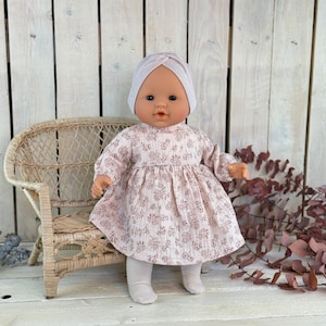May include: A doll wearing a pink floral dress and a light pink turban. The doll is sitting on a wicker chair.