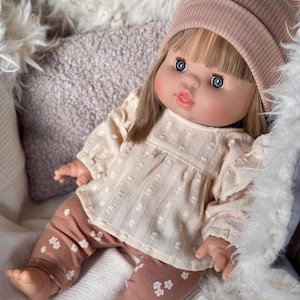 Könnte beinhalten: Puppe mit blauen Augen, trägt eine hellrosa Mütze, eine cremefarbene Bluse mit weißen Punkten und braune Leggings mit weißen Blumenmustern. Die Puppe liegt auf einer weichen, strukturierten Oberfläche.