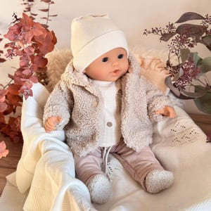 Könnte beinhalten: Eine Babypuppe mit cremefarbener Mütze, beiger Sherpa-Jacke, weißem Hemd und rosa Hose. Die Puppe sitzt in einem mit einer weißen Decke ausgekleideten Korb, mit getrockneten floralen Akzenten im Hintergrund.
