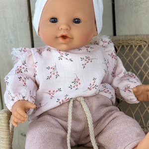 Könnte beinhalten: Eine Babypuppe mit blauen Augen und einer weißen Mütze, bekleidet mit einem hellrosa Langarmshirt mit kleinem rot-grünem Blumenmuster und Rüschen an den Schultern, kombiniert mit einer malvenfarbenen Strickhose mit weißem Kordelzug.