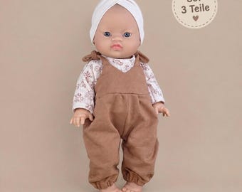 May include: A doll wearing a white turban, a long-sleeved shirt with a brown floral pattern, and brown overalls. The doll has blue eyes and a neutral expression. A circular tag in the upper right corner displays the text Set 3 Teile.