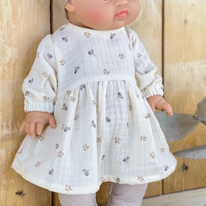 May include: A doll wearing a long-sleeved, off-white dress made of textured fabric. The dress features a small, repeating floral pattern in muted blue and brown tones, a gathered waistline, and elasticized cuffs on the sleeves. The doll has light skin.