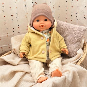 Può includere: Una bambola vestita con una giacca con cappuccio giallo chiaro, una camicia con stampa floreale e pantaloni color crema. La bambola ha anche un berretto color talpa con orecchie da gatto. È seduta in un cestino intrecciato su una coperta color crema.