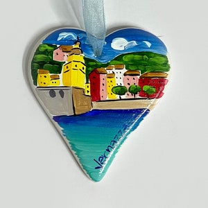 May include: A heart-shaped ceramic ornament featuring a painted scene of a coastal town. The town is depicted in shades of yellow, pink, and red, with a blue sea and green hills in the background. The ornament has a light blue ribbon attached.