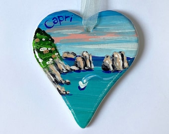 May include: A heart-shaped ceramic ornament featuring a hand-painted coastal scene of Capri with blue water, rocky cliffs, and green foliage. The word Capri is written in blue at the top. A light blue ribbon is attached for hanging the ornament.