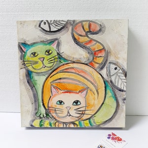 Könnte beinhalten: Ein Aquarellbild von zwei Katzen auf weißem Hintergrund. Die Katze im Vordergrund ist orange und weiß mit grünen Streifen. Die Katze im Hintergrund ist grün und gelb mit schwarzen Streifen. Beide Katzen haben schwarze Augen und Schnurrhaare.