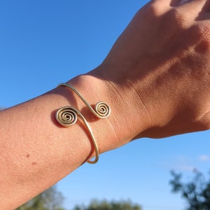 Op de afbeelding: Een gouden metalen armband met twee spiraalontwerpen. De armband is open en verstelbaar.
