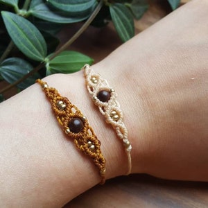 Könnte beinhalten: Zwei Makramee-Armbänder, eines in warmem Braun und das andere in Creme, jeweils mit einer zentralen dunkelbraunen Perle und kleinen goldfarbenen Perlen. Die Armbänder werden am Handgelenk einer Person präsentiert und zeigen die komplizierte Knotenarbeit.