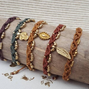 Könnte beinhalten: Fünf geflochtene Armbänder mit goldenen Perlen und Anhängern. Die Anhänger umfassen ein Blatt, eine Träne und ein Wirbeldesign. Die Armbänder sind in Braun-, Grün- und Lilatönen gehalten.