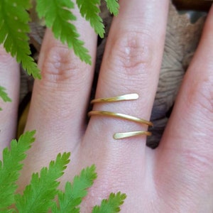 Könnte beinhalten: Ein goldfarbener, doppelt gewickelter Ring an einem Finger. Der Ring ist aus dünnem, glattem Metall gefertigt und hat ein schlichtes, minimalistisches Design.