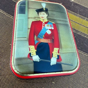 May include: A rectangular tin with a portrait of a person in a red military uniform with a blue sash and medals. The tin has a red border and a slightly distressed appearance, suggesting age.