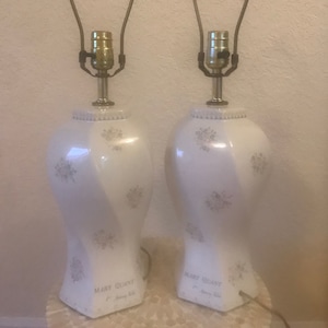 Könnte beinhalten: Zwei weiße Keramik-Tischlampen mit Blumendesigns und goldenen Akzenten. Die Lampen haben ein Mary Quant-Etikett und sind für Standard-Glühbirnen verdrahtet.