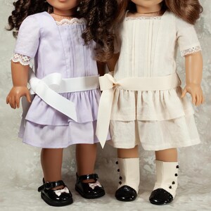 May include: Two dolls wearing matching dresses. One doll is in a light purple dress with a white ribbon belt and black shoes. The other doll is in an off-white dress with a cream ribbon belt and black and white boots.
